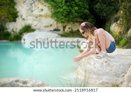 Young girl playing by natural swimming pool in Bagno Vignoni, with thermal spring water and calcium carbonate deposits, which form white concretions and waterfall. Tuscany, Italy. Royalty-Free Stock Photo #2118916817