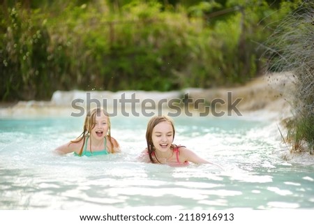 Two young sisters bathing in natural swimming pool in Bagno Vignoni, with thermal spring water and calcium carbonate deposits, which form white concretions and waterfall. Tuscany, Italy. Royalty-Free Stock Photo #2118916913