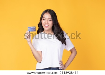 Portrait of a happy young asian woman wearing white shirt holding bank card, credit card isolated on yellow background. Business online shopping concept.