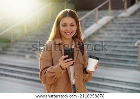 Enchanting blonde young woman waiting for phone message while carrying coffee cup on the street. Stylish girl in spring clothing holding smartphone and take-away cappuccino. Royalty-Free Stock Photo #2118958676