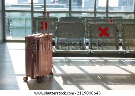 Travel pink suitcase or carry-on bag near empty waiting chairs in airport terminal during COVID-19 omicron pandemic with social distancing signs on chairs. Transportation travel in new normal.