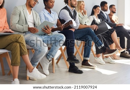 Crop image of diverse employees sit on chairs in row listen to seminar or training in office. Job candidates or applicants in line wait for interview for vacant position. Employment, hiring concept. Royalty-Free Stock Photo #2119007924
