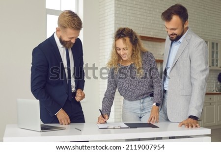 Happy young married couple buying new house or apartment. Smiling boyfriend and girlfriend standing at table in kitchen of new home and together signing contract given by real estate agent or realtor