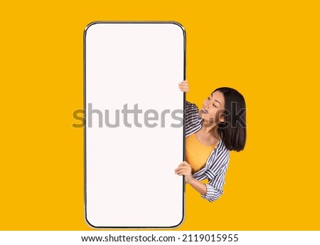 Great Offer. Portrait of excited asian woman peeking out big giant vertical cell phone with white blank screen and looking at device display. Gadget with empty free space mock up, yellow orange wall