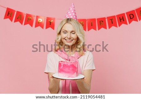 Elderly happy smiling woman 50s with closed eyes wear t-shirt birthday hat hold cake with candle making wish isolated on plain pastel pink background studio portrait. Celebration party holiday concept