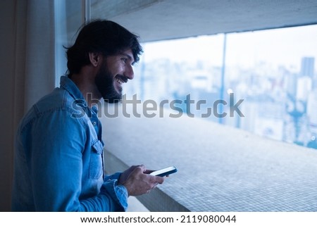Side view of young man using a smartphone at night time on his balcony with city view landscape in the background. Mobile phone, technology, concept. Texting on the phone. High quality photo