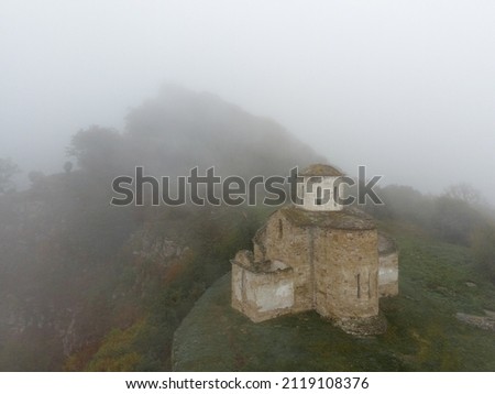 Aerial view of ancient Christian Sentinel Church, Caucasus. It is located on spur of mountain range. Thick fog covers white stone walls of church and greenery around. Roof is covered in moss Royalty-Free Stock Photo #2119108376