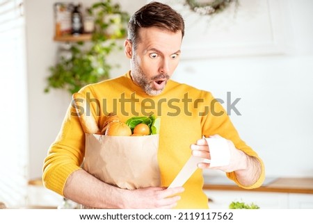 Surprised man looking at store receipt after shopping, holding a paper bag with healthy food. Guy in the kitchen. Real people expression. Inflation concept. Royalty-Free Stock Photo #2119117562