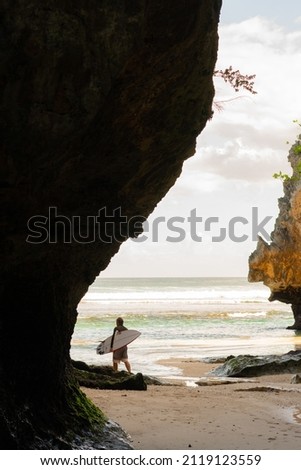 A view of the beach with rocks and a young man in a swimming suit walking on the sandy shore with a surfboard.