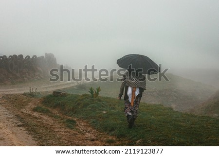 Woman walking alone with umbrella on muddy pasture in foggy, rainy day.  Back view