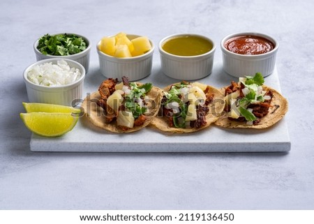 Traditional Mexican street food called "Tacos al pastor" prepared with pork, tortillas, pineapple, cilantro, onion, green sauce, red sauce and lemons. On white texture background.
