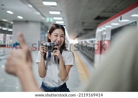 Young beautiful Asian female tourist taking portrait photo with film camera with friend, smile and enjoyment at train station platform, happy travel lifestyle by subway transport vacation trip.