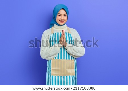 Cheerful young Asian Muslim woman wearing hijab and apron, open sign to greet and greet customers isolated on purple background. People housewife muslim lifestyle concept Royalty-Free Stock Photo #2119188080
