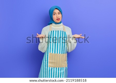 Confused young housewife woman in hijab and striped apron, shrugging shoulders look puzzled have no idea nothing to say isolated on purple background. People housewife muslim lifestyle concept
