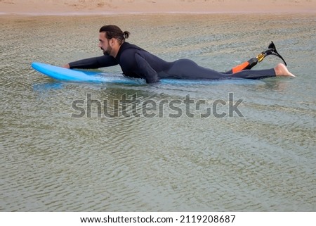 Man with beard and curly hair swimming. Mid adult man in surfing suit getting ready to surf in water, lying on surfboard. Sport, leisure, disability, active lifestyle concept
