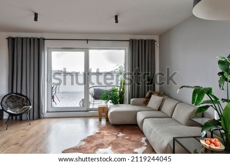 Modern living room in stylish interior with large window. Comfortable sofa and armchair on wooden floor and green plant in the room in luxurious apartment. Royalty-Free Stock Photo #2119244045