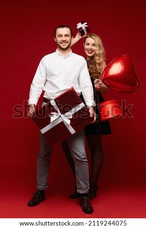 A lovely heterosexual couple posing with gift boxes and a red balloon in the shape of a heart, a pretty blonde woman and a handsome young man posing in the red background