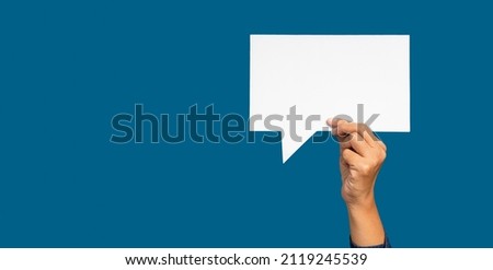 A speech bubble concept. Hand holding of an empty white speech bubble against a blue background. Space for text. Close-up photo.