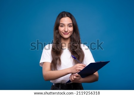 Attractive Caucasian or Arab young woman in a casual white t-shirt smiling looking at the camera, writing on a clipboard and taking notes isolated on a blue studio background. Concept business, work.