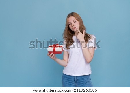 Romantic gift. Happy young woman opens valentine's day gift on blue studio background, copy space. Beautiful woman celebrating a romantic holiday, holding a box with a red ribbon in a white t-shirt