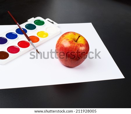 Watercolor set with brush and red apple on the white paper sheet over the black surface
