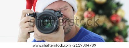 Man in red santa hat taking pictures with camera near christmas tree. New year photo session concept