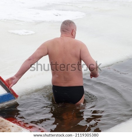 Elderly Caucasian man walks into the icy water from wooden Pier in an ice hole in winter.