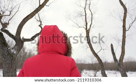 Rear view image of adult woman dressed in red winter jacket walking away from camera in snowy cold winter blurry landscape background