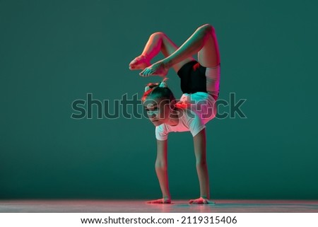 Graceful handstand. Little flexible girl, rhythmic gymnastics artist training isolated on green studio background in neon pink light. Doing exercises in flexibility. Beauty, sport, challenges