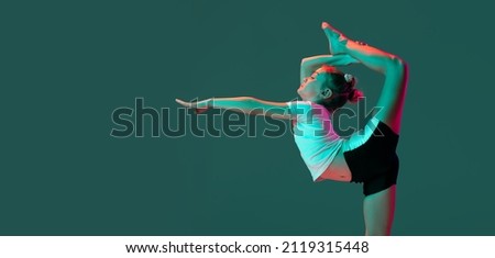 Little flexible girl, rhythmic gymnastics artist training isolated on green studio background in neon pink light. Doing exercises in flexibility. Beauty, sport, challenges, studying, ad