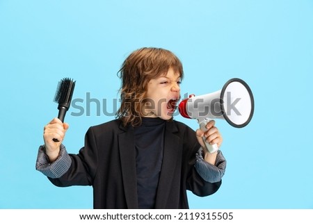 Pay attention, news. Liitle cute boy, kid in image of stylish hairdresser, barber shouting at megaphone isolated on blue background. Concept of occupation, emotions, childhood, studying