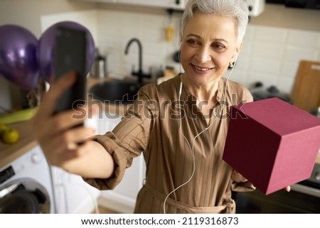 Fashionable stylish beautiful woman in lovely dress and earphones holding pink box of her birthday present and phone to make selfie, standing against kitchen, having cute smile. Human and technology
