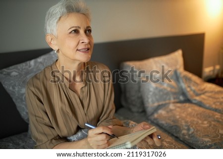 Mature pretty stylish Caucasian female with short grey hair looking up while sitting in bed, with pensive face expression, making notes, planning week, or writing down in diary before going to sleep Royalty-Free Stock Photo #2119317026