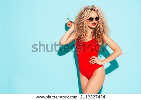 Young beautiful smiling woman posing near blue wall in studio.Sexy model in red swimwear bathing suit.Positive female with curls hairstyle. Holding penny skateboard.Happy and cheerful Royalty-Free Stock Photo #2119327454