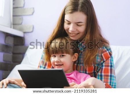 Two happy cute sisters are sitting on the couch and watching cartoons together on a tablet computer. The older sister shows the funny younger sister educational games online on the tablet screen.