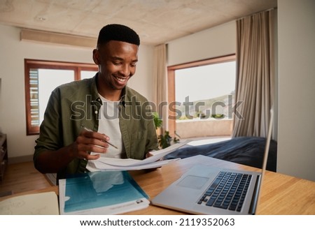 Young adult black male smiling while studying with paper notes, book and laptop in modern apartment.  Royalty-Free Stock Photo #2119352063