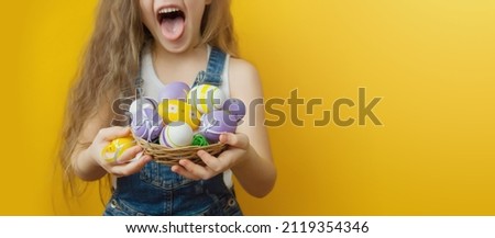 A mischievous girl in a white T-shirt and denim overalls holds a small wicker basket with Easter eggs in her hands on a yellow background. Selective focus. Close-up. Copyspace. Layout for advertising.