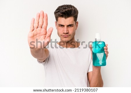 Young caucasian man holding mouthwash isolated on white background standing with outstretched hand showing stop sign, preventing you.