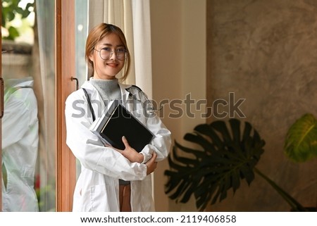 A portrait of a pretty Asian doctor in white coat standing in the office with confident smile carrying 
 books. for medical and health care concept.