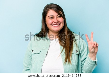 Young caucasian overweight woman isolated on blue background joyful and carefree showing a peace symbol with fingers.