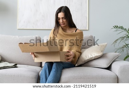 Angry confused woman unpacking parcel, wrong or broken online store order, sitting on couch at home, dissatisfied female looking in cardboard box, bad delivery service, displeased by post shipping Royalty-Free Stock Photo #2119421519