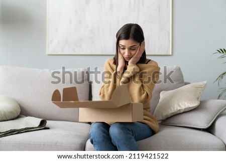 Angry confused woman unpacking parcel, wrong or broken online store order, sitting on couch at home, dissatisfied female looking in cardboard box, bad delivery service, displeased by post shipping Royalty-Free Stock Photo #2119421522
