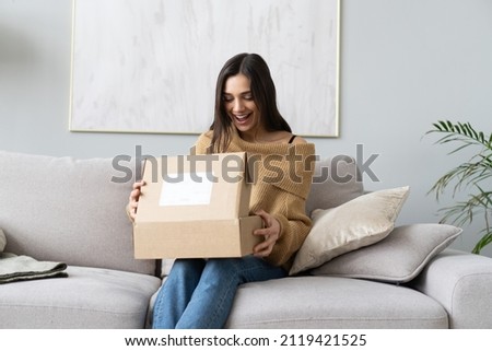 Happy young woman sit on couch in living room unpack cardboard box buying goods on Internet, smiling excited millennial girl open carton parcel order, shopping online, good delivery concept Royalty-Free Stock Photo #2119421525