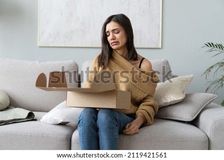 Angry confused woman unpacking parcel, wrong or broken online store order, sitting on couch at home, dissatisfied female looking in cardboard box, bad delivery service, displeased by post shipping Royalty-Free Stock Photo #2119421561