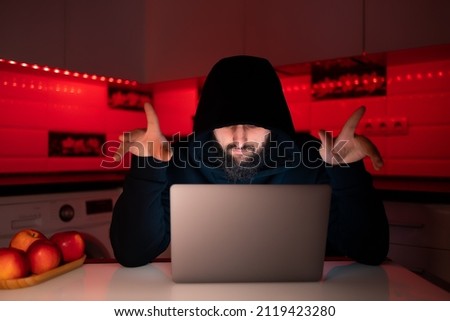 a hacker in a black hoodie with a hood on his head is sitting at a laptop showing his thumbs up rejoicing at a successful hack