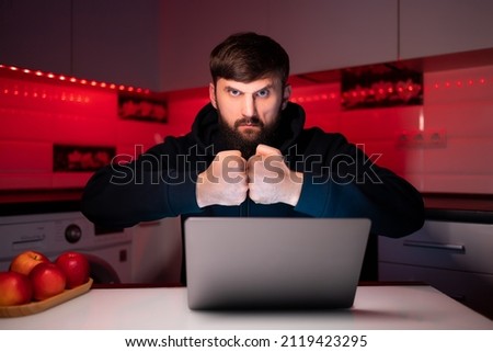a hacker in a black hoodie is sitting at a laptop with his fists together in front of him