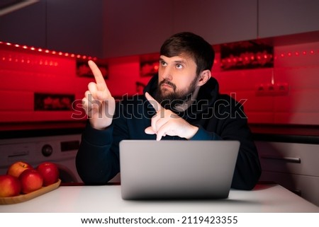 a hacker in a black hoodie is sitting at a laptop pointing his fingers to the side