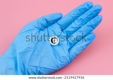 A hand in a medical glove holds a pill with a Cedi sign on a pink background. Expensive medicine in Ghana. Pharmaceutical business.