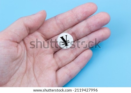 A hand holds a medical pill with an yen or yuan currency sign on a blue background. The concept of a rise in the price of medicines in Japan or China. Pharmaceutical business.