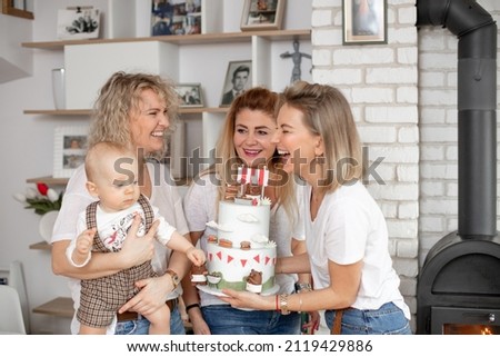 Portrait of delightful laughing family three women congratulating baby looking at cake wearing square overall, celebrating birthday party. Presents and gifts with cake, decorated by bears cartoons.